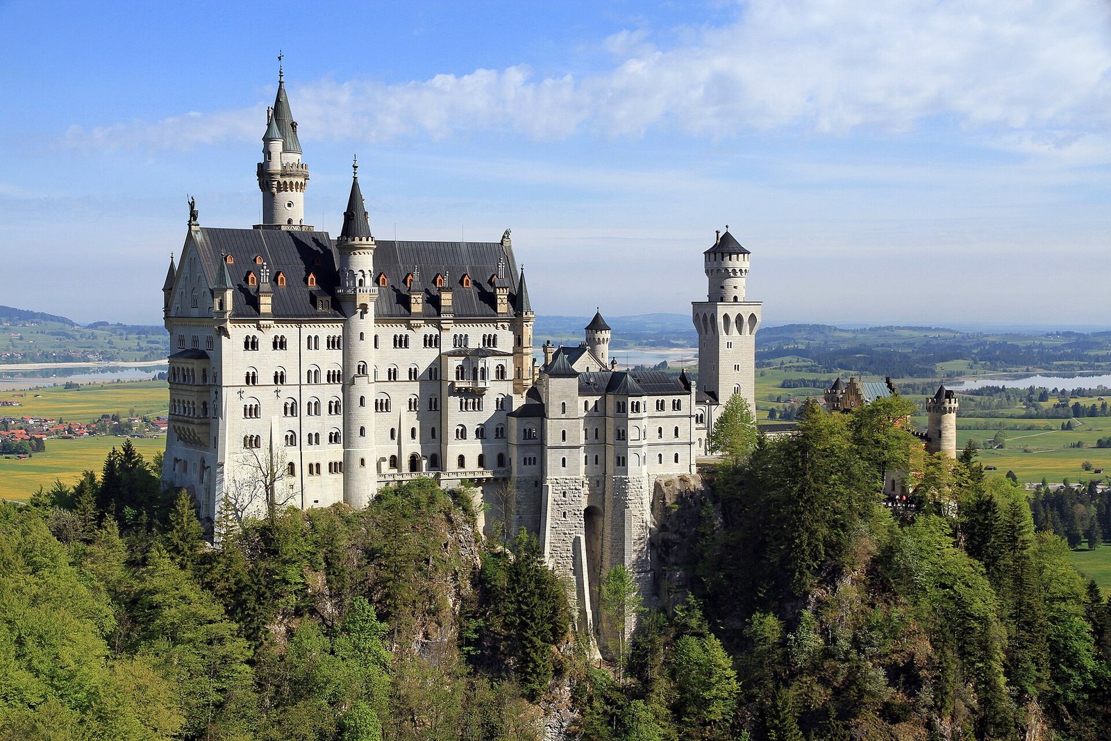 A white castle with slate roofs, turrets and a round tower on a wooded crag above green fields and lakes, under a blue sky with thin cloud