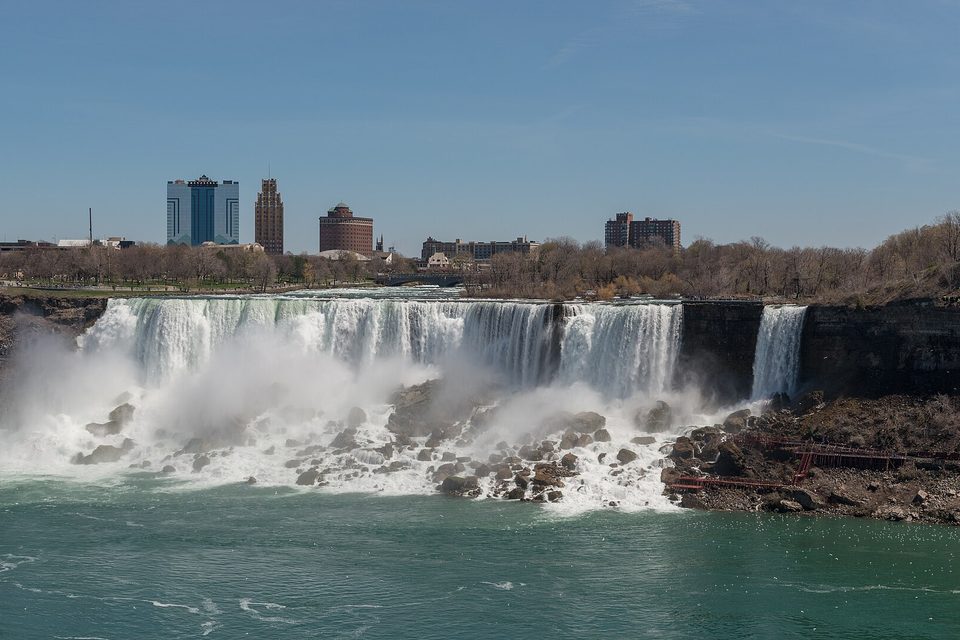 A broad low waterfall and a narrow one beside it dropping over a rocky ledge, with the towers of Niagara Falls, New York, on the skyline