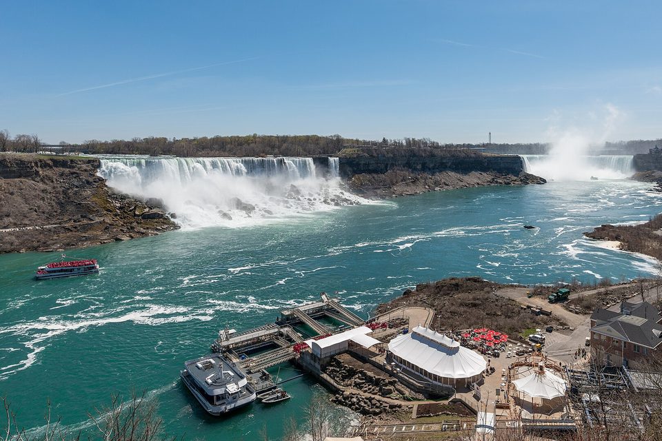 A turquoise river gorge with a wide curved waterfall in the distance and a boat landing and viewing decks on the near shore