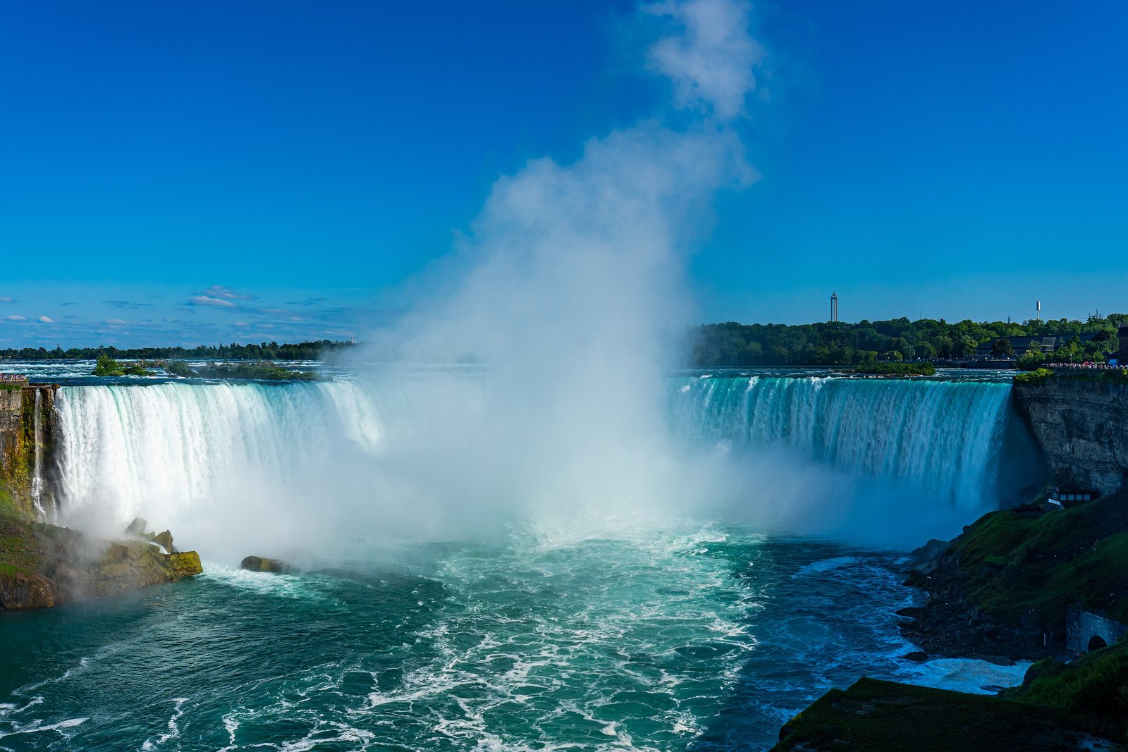 A curved wall of green and white water falling into a turquoise pool, a tall column of mist rising against a clear blue sky