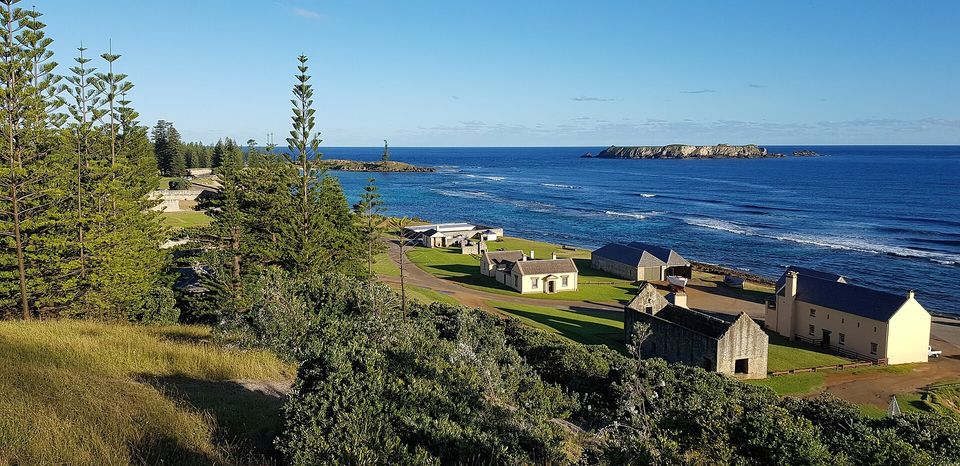 From a hillside of tall pines, a cluster of low cream and grey stone buildings stands on a green point beside a deep-blue, surf-lined sea, with a rocky islet offshore