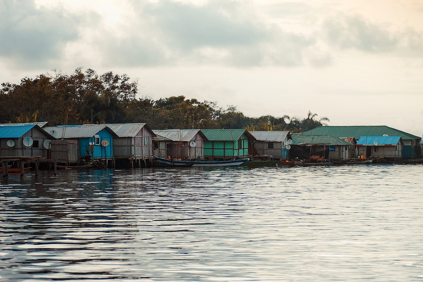 A row of painted stilt houses with satellite dishes across a rippled lake in soft evening light, a boat moored at the right and forest behind