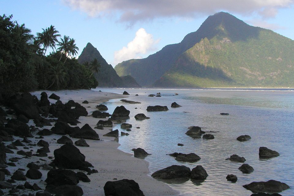 Black boulders scattered along a pale beach and shallow lagoon, below steep green mountains in soft light