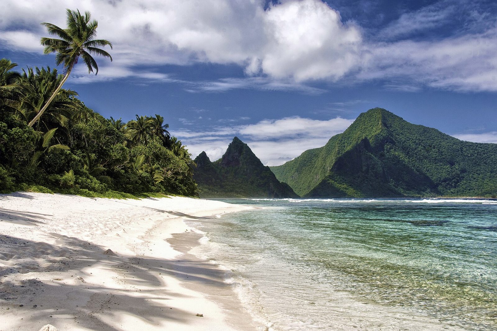 A white beach under leaning palms, clear shallow water, and Olosega's two green peaks across the strait