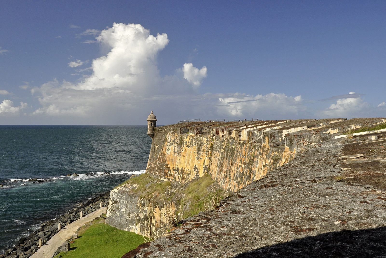 Sun-washed stone fortress walls stepping down to a rocky shore and surf, a sentry box on a corner