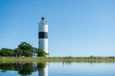 A tall white lighthouse with a black band, reflected in calm water beside a red cottage and trees