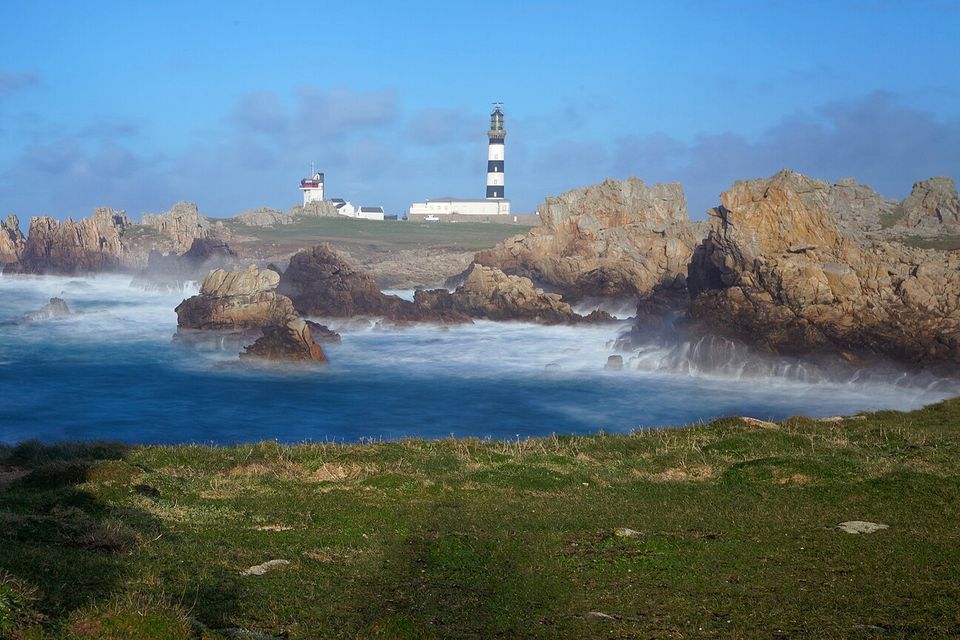A black-and-white striped lighthouse and low white buildings stand on a green headland above jagged golden rocks and misty, wave-blurred sea