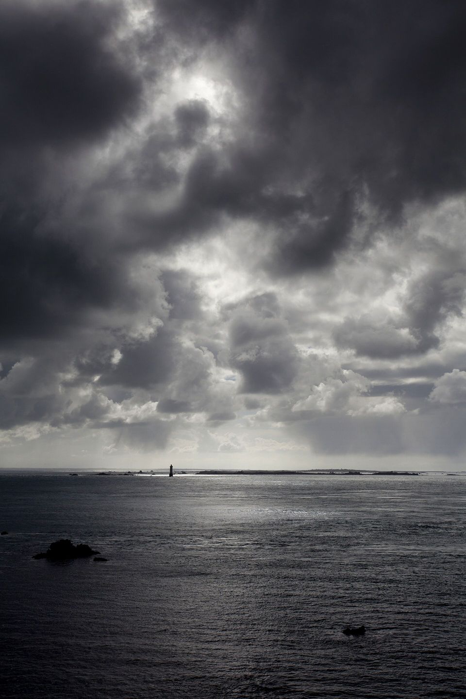 Under a towering sky of dark storm cloud, a lone lighthouse stands as a tiny silhouette on a silver sea, low islands on the horizon