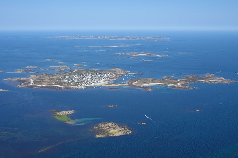 From high above a deep-blue sea, a small low island packed with grey village roofs is ringed by pale sand and scattered rocky islets, with a longer island on the far horizon