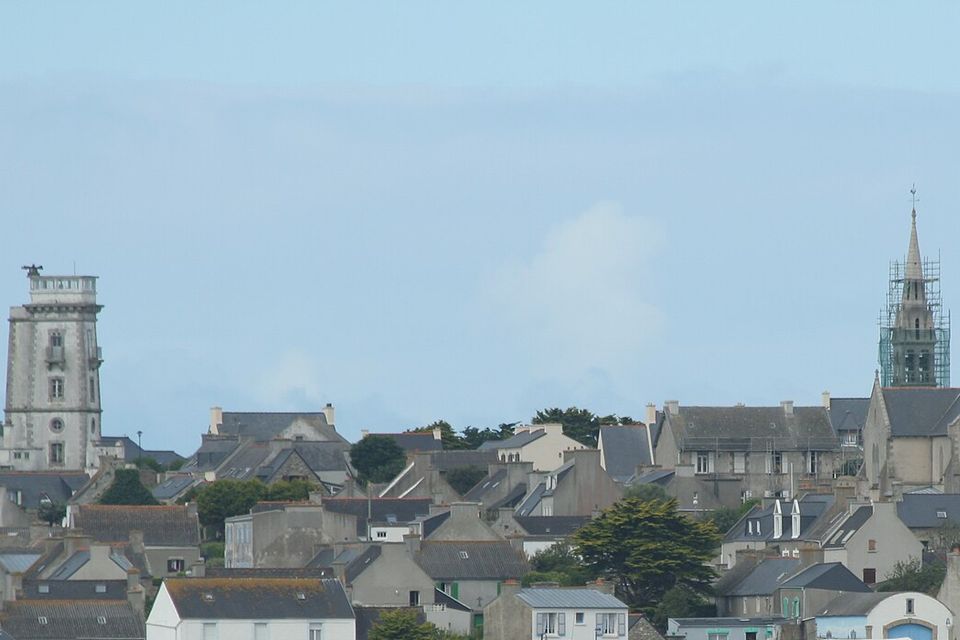 A tight village of grey stone houses with slate roofs fills a low island, a square signal tower at left and a church with a scaffolded spire at centre, under a pale blue sky