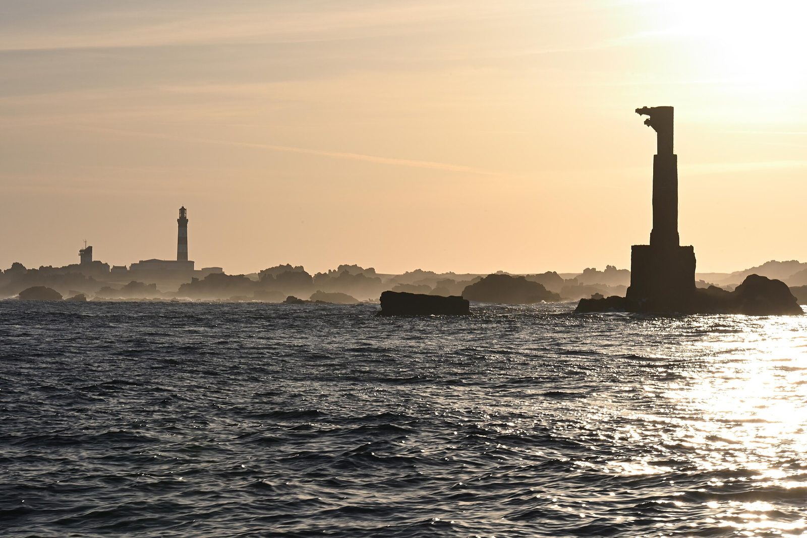 At golden hour a dark tower stands on a reef in a sparkling sea, and the slim Créac'h lighthouse rises on the low skyline of Ouessant under a soft gold sky
