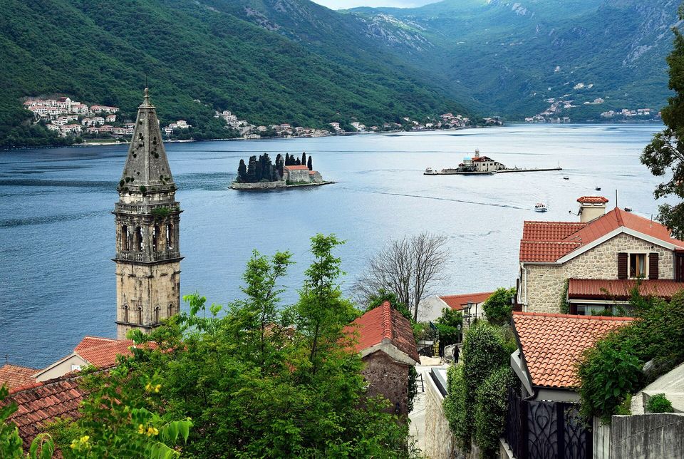 A blue bay with two small islets, one dark with cypresses and one holding a domed church, seen past a tall stone bell tower and red roofs in the foreground, forested mountains behind
