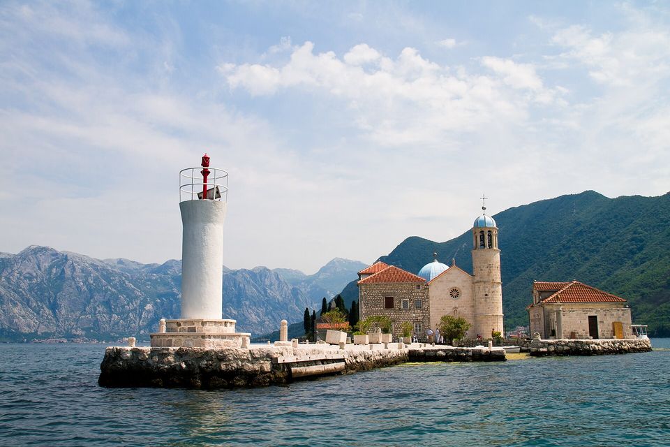 A white lighthouse with a red lamp at the end of a low stone quay, a small church with a pale blue dome and a bell tower beside it, mountains beyond and blue water in front