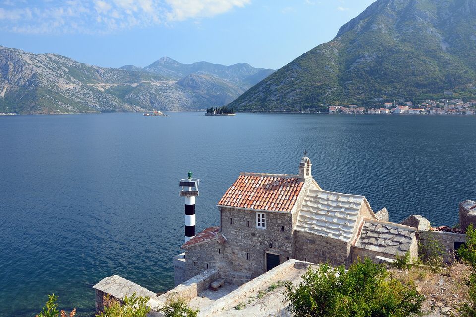 A wide blue bay between steep mountains seen from a hillside, a small stone chapel beside a black-and-white striped light in the foreground, two tiny islets far off and a town on the right shore