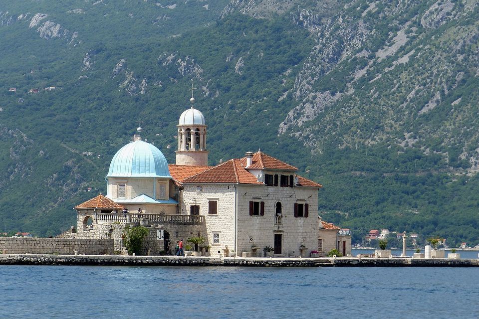 A stone church with a pale blue dome and a slim bell tower with a small cupola on a low islet, steep green mountains behind and two tiny visitors at the left of the quay