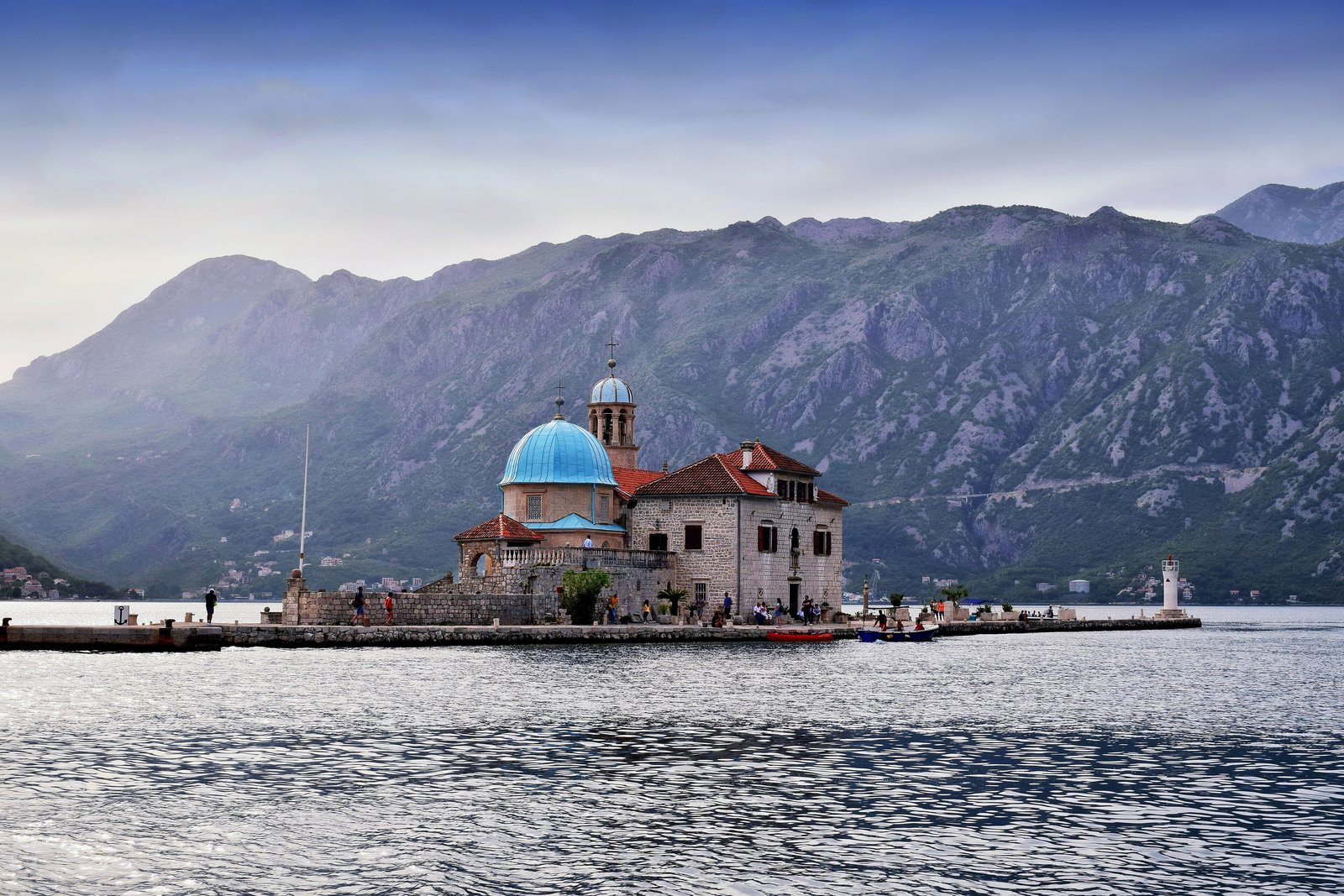 A stone church with a pale blue dome and a bell tower on a small flat islet with a stone quay and a white lighthouse at the right, steep purple-grey mountains behind and rippled water in front