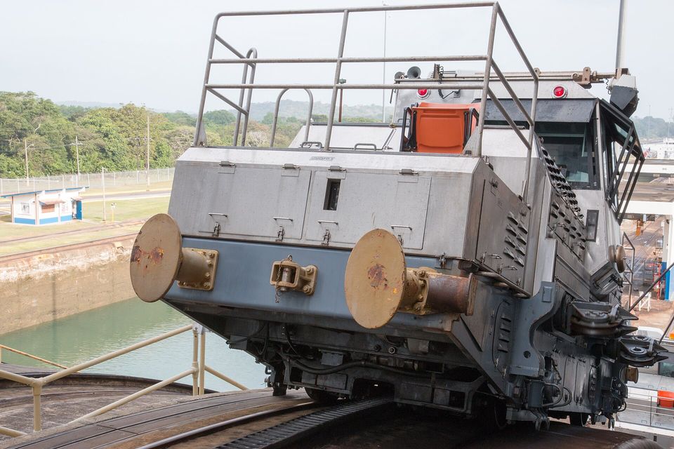 The front of a grey and blue electric locomotive with two rusty round buffers and a railing, an empty orange seat above, on a track beside a green lock chamber