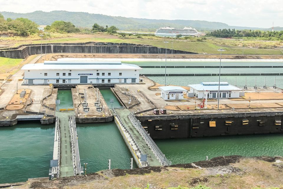 A modern lock complex with white control buildings and green water in the chambers, a cruise ship far behind on the horizon