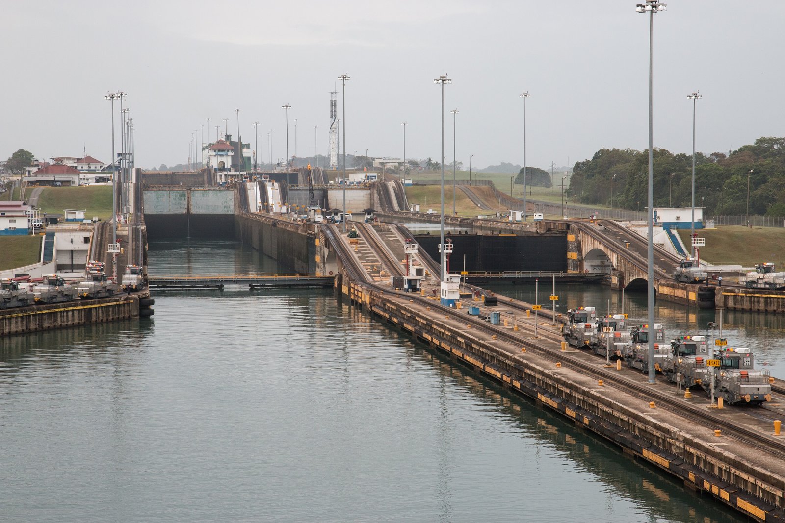 A flight of concrete lock chambers stepping away from the camera over still water, a row of small electric locomotives on the centre wall at right and tall lamp posts along the walls, under a grey sky