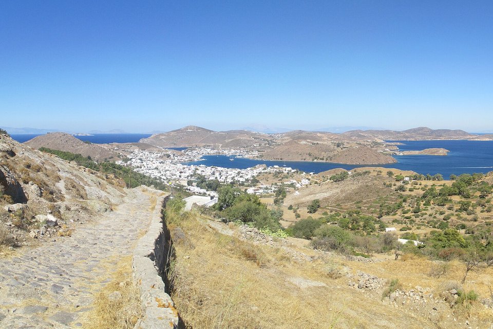 A cobbled path edged by low stone walls running down a dry hillside of olive trees toward a white harbour town on a deep blue bay
