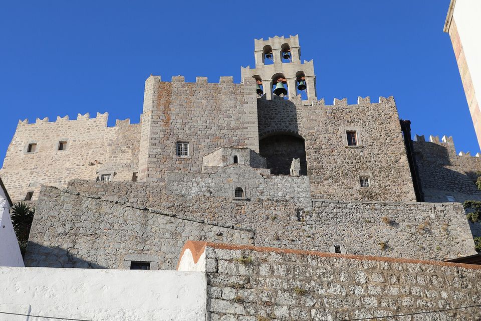 The battlemented grey stone walls of a monastery rising in tiers, with a triple bell-gable on top, seen from below against a deep blue sky