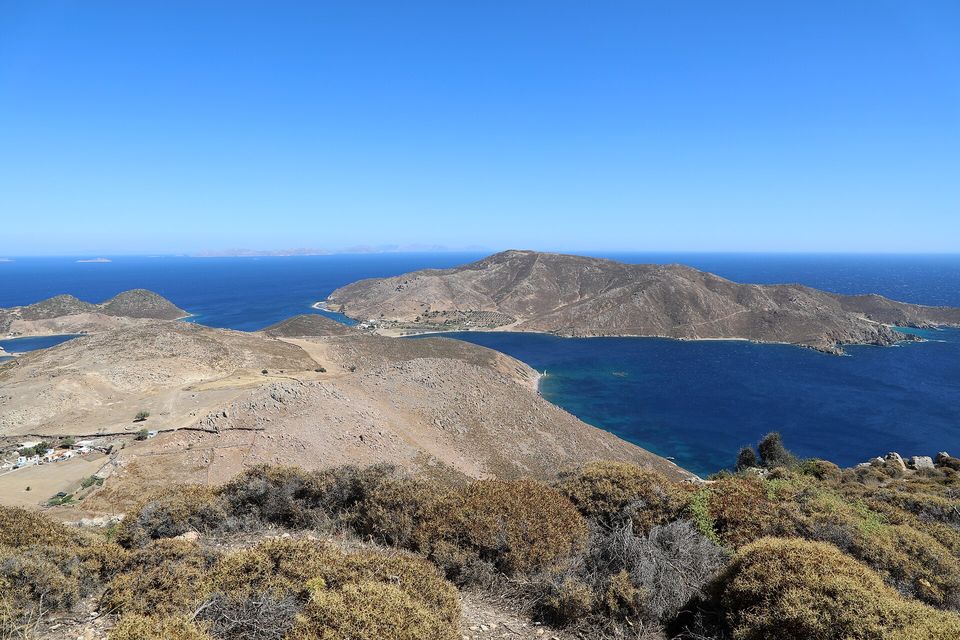 A bare brown headland running out to a small island in deep blue water under a clear sky, low grey scrub in the foreground