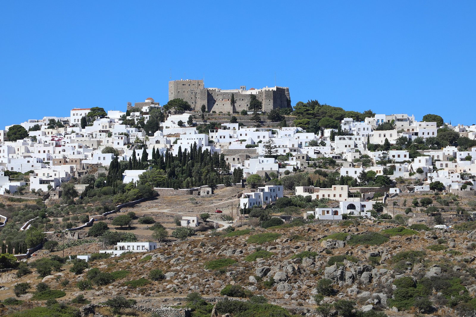 A white hill town spread across a rocky slope under a blue sky, the dark crenellated walls of a fortress-like monastery on the summit