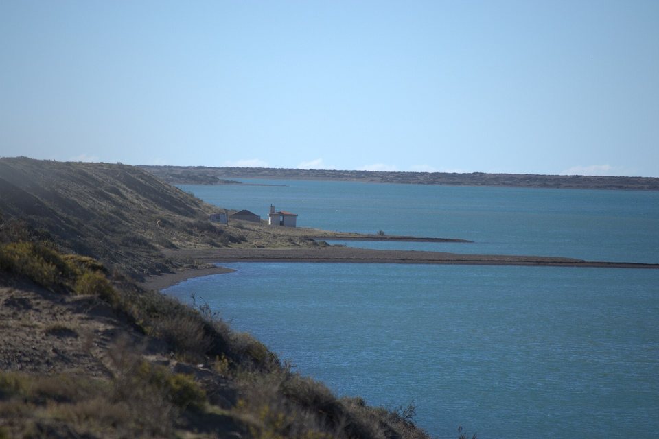 A long calm blue lagoon behind a low shingle spit, a small building on the shore below a scrubby slope