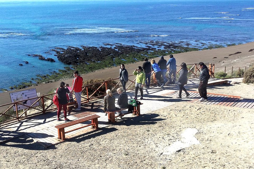 Visitors at a wooden railing on a sandy bluff looking down at a grey beach, a dark rock reef and the open Atlantic