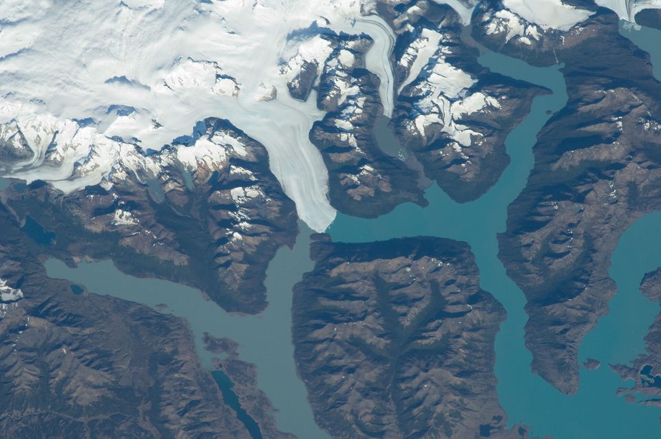 From orbit, a white tongue of ice flowing from a snowfield between brown mountains into branching arms of a blue-green lake