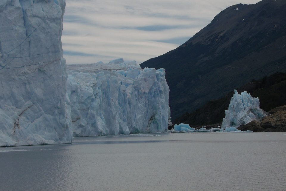 Open grey water between a tall ice cliff on the left and a dark rocky shore on the right, a leftover block of ice against the rocks