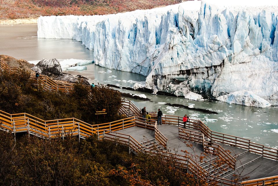 Zig-zagging walkways with orange railings descending through autumn shrubs toward a channel and a towering blue-white ice wall
