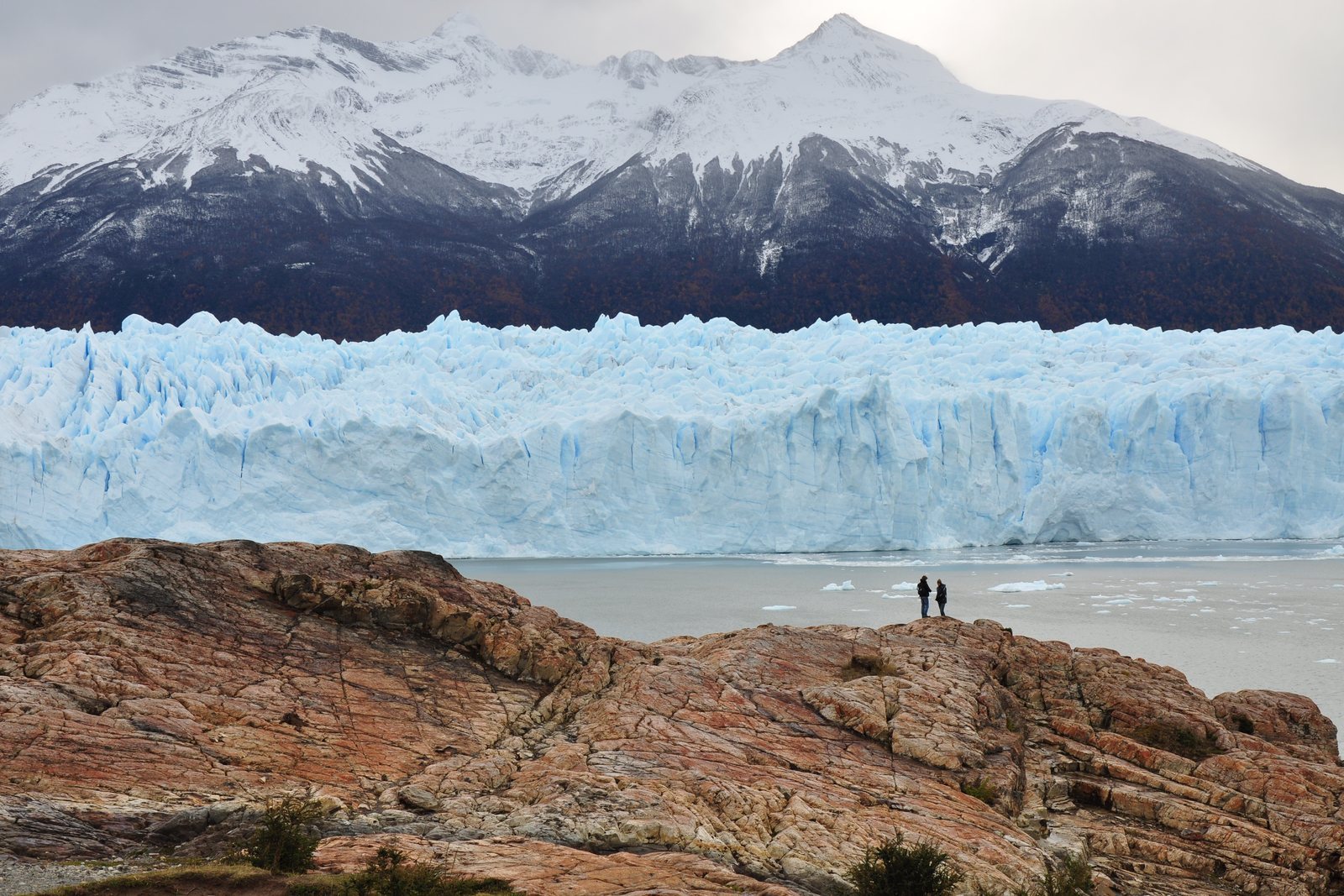 A glacier front stretching across the frame as a wall of pale blue ice, a snow-covered mountain behind, two people tiny on a rock outcrop