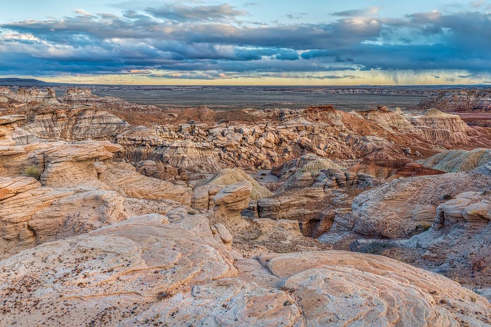 A wide view of eroded tan and blue badland ridges under heavy clouds with a band of light on the horizon
