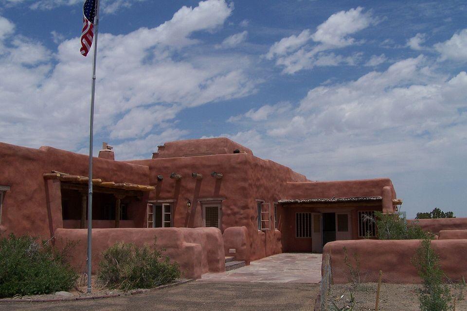 A low adobe-style building with a flagpole and a paved entrance terrace under a cloudy blue sky