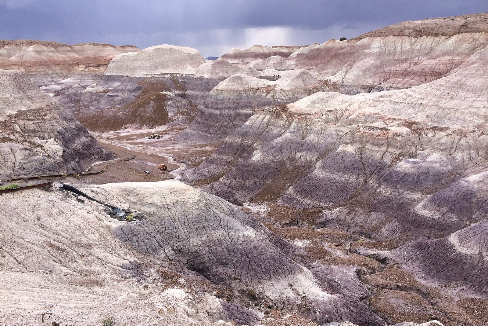 Grey, blue and purple banded clay hills cut by gullies, under a dark sky with rain falling in the distance