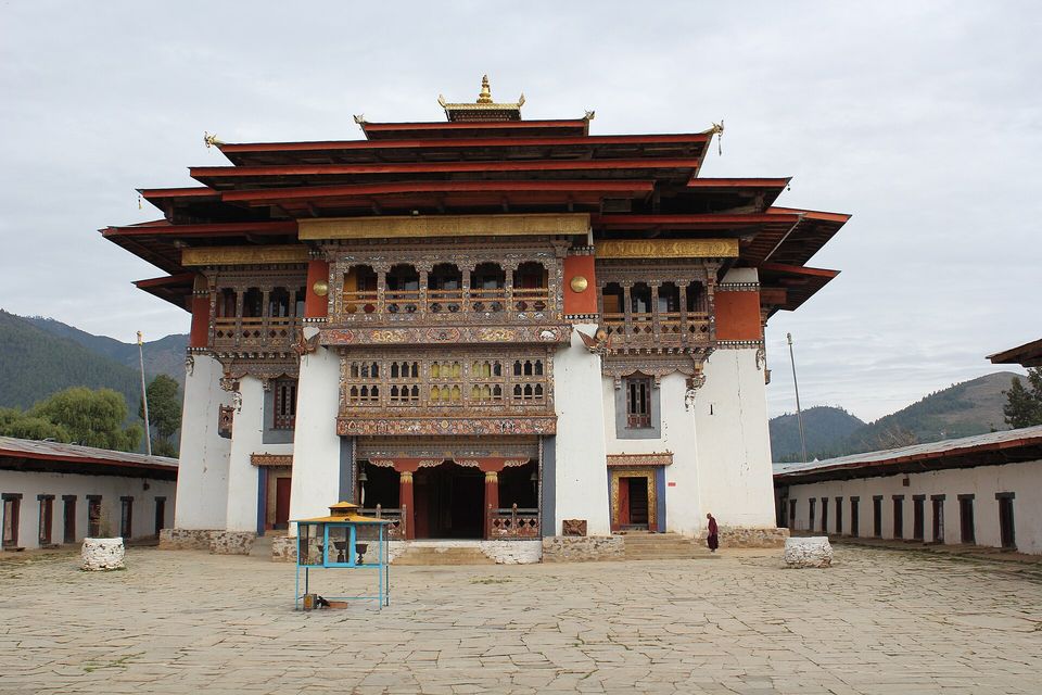 A tall whitewashed monastery with tiered red roofs, carved wooden windows and a gilded finial stands across a stone-paved courtyard, green hills behind