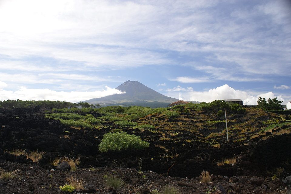 The Pico volcano in the distance over black lava ground and low scrub, cloud around its base