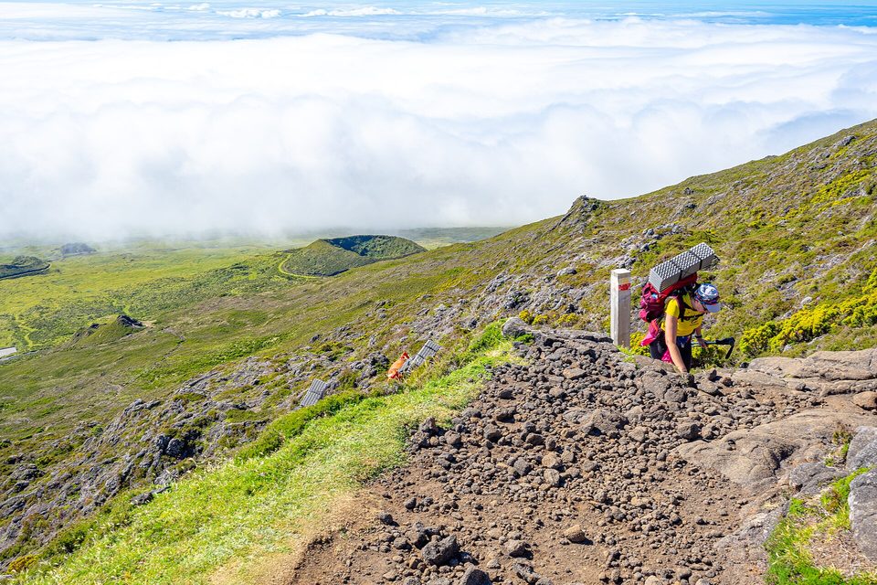 A hiker on the summit trail beside a route post, a sea of cloud below