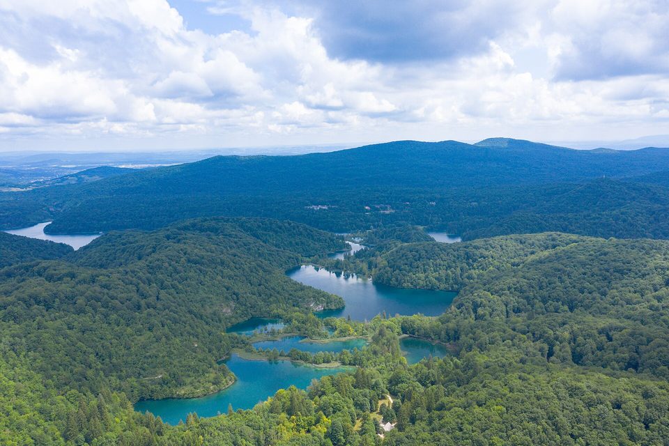 A chain of green and turquoise lakes stepping down through dense forest, seen from the air under broken cloud