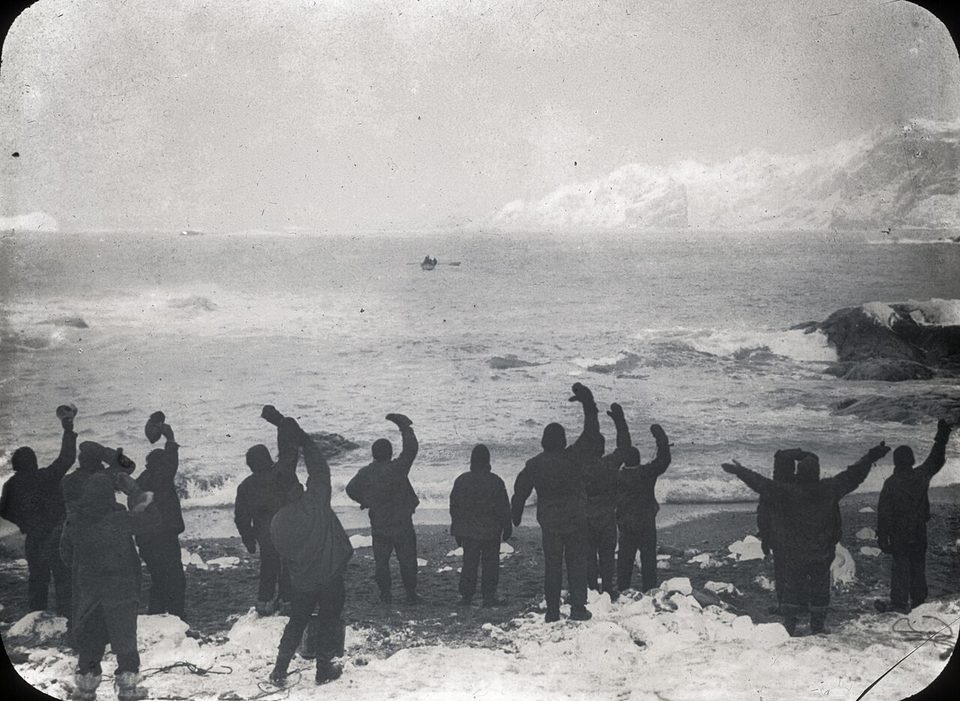 A black-and-white photograph of men in dark clothing on a snowy shore waving toward a distant boat on a rough sea