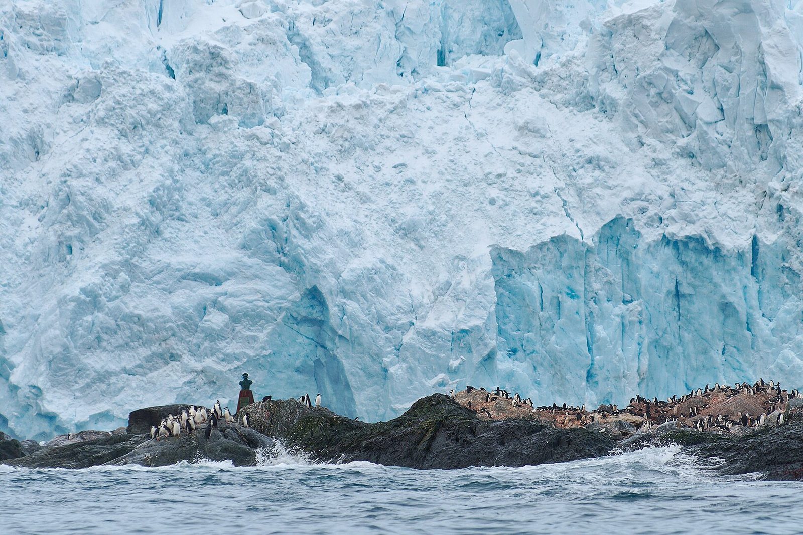 A small stone bust on a rock with chinstrap penguins before a blue glacier face