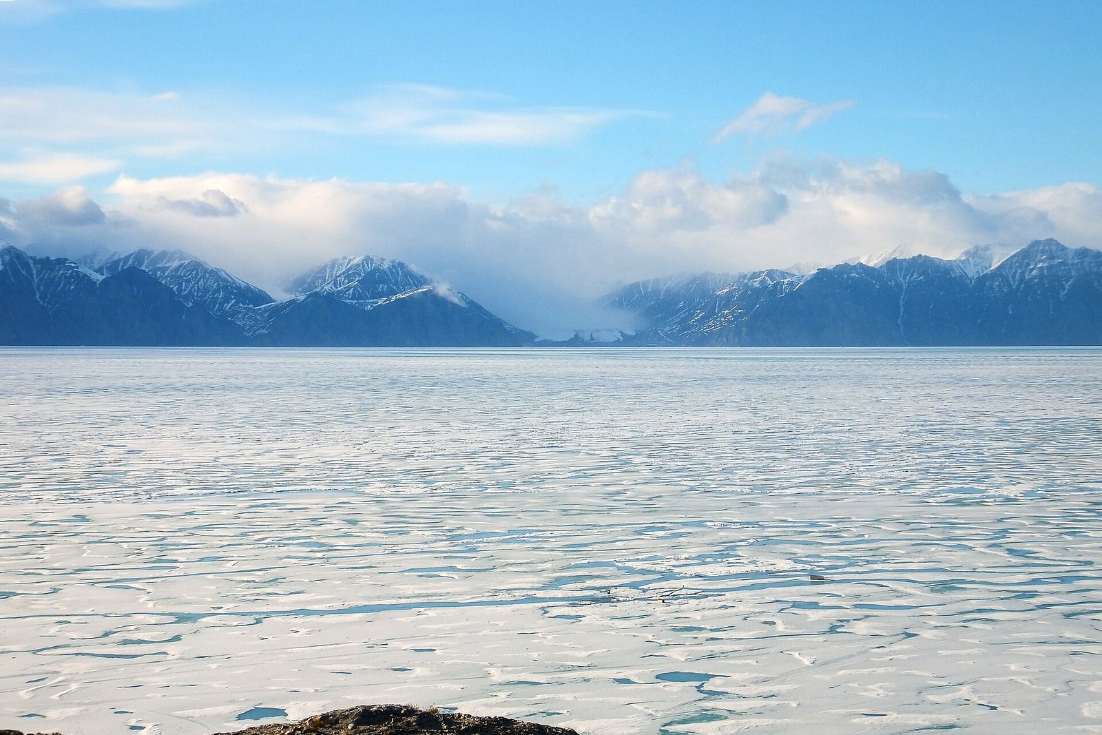 Snow-streaked peaks under cloud across a wide sound of sea ice patterned with meltwater pools