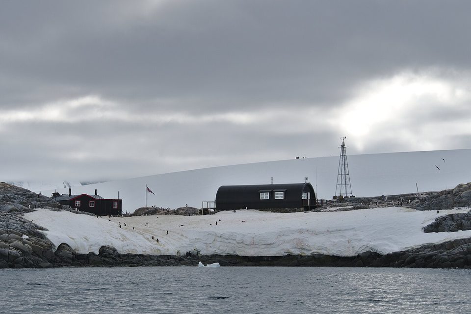 A low rocky island seen from the sea, with a red-trimmed hut, a black curved Nissen hut, a flag and a radio mast on snow under grey cloud