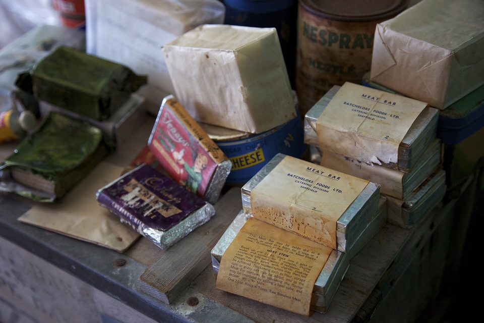 Stacked tins and paper-wrapped blocks labelled "Meat Bar", a cheese tin and chocolate bars on a worn wooden shelf
