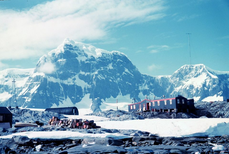 A faded colour photo of the red-trimmed huts and a Nissen hut on snowy rocks, with fuel drums, below a great snow-capped mountain