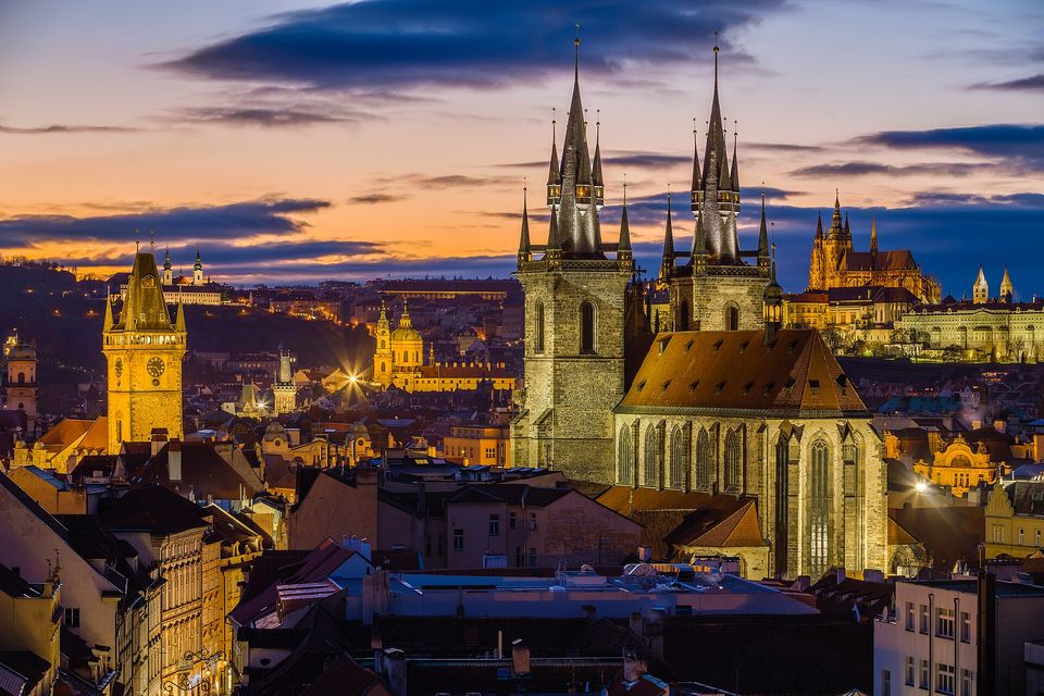 A city of red-tiled roofs at dusk under an orange and blue sky, a golden tower at the left, two dark Gothic spires at the centre and a lit hilltop castle in the distance