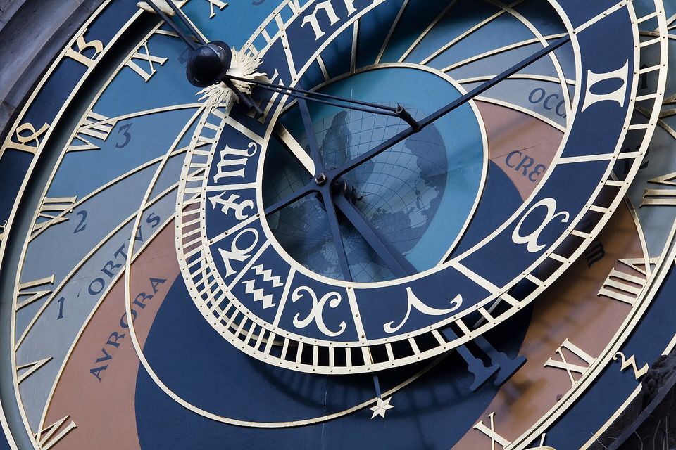 A close view of an astronomical clock dial with gilt Roman numerals and zodiac signs on blue, dark iron hands crossing over a map-like disc