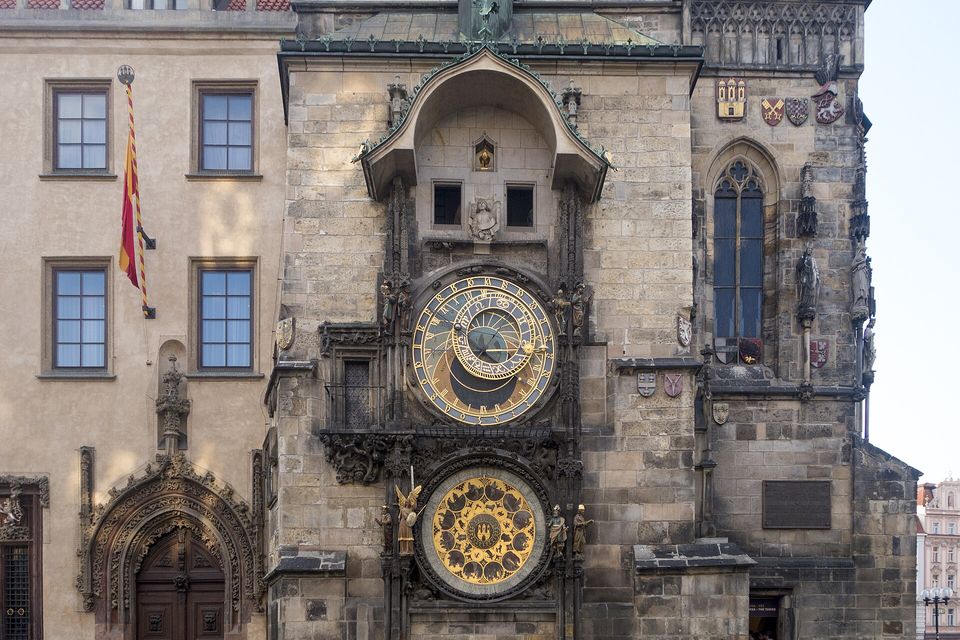 A stone tower wall with a hooded balcony window at the top, a round astronomical clock dial in the middle and a round gold calendar dial below, a striped flag at the left
