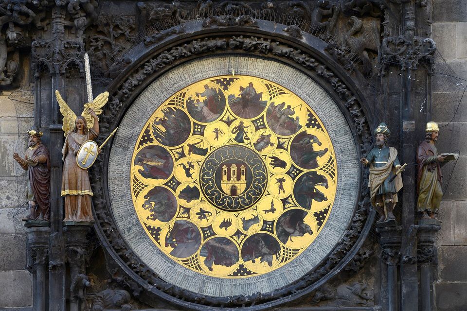 A round gold calendar dial painted with small scenes of the months around a central city emblem, an angel with a sword and shield at the left and two robed stone figures at the right, on dark carved stonework